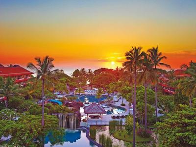 Vista al atardecer de un resort tropical con piscina, palmeras y techo rojo junto al mar.