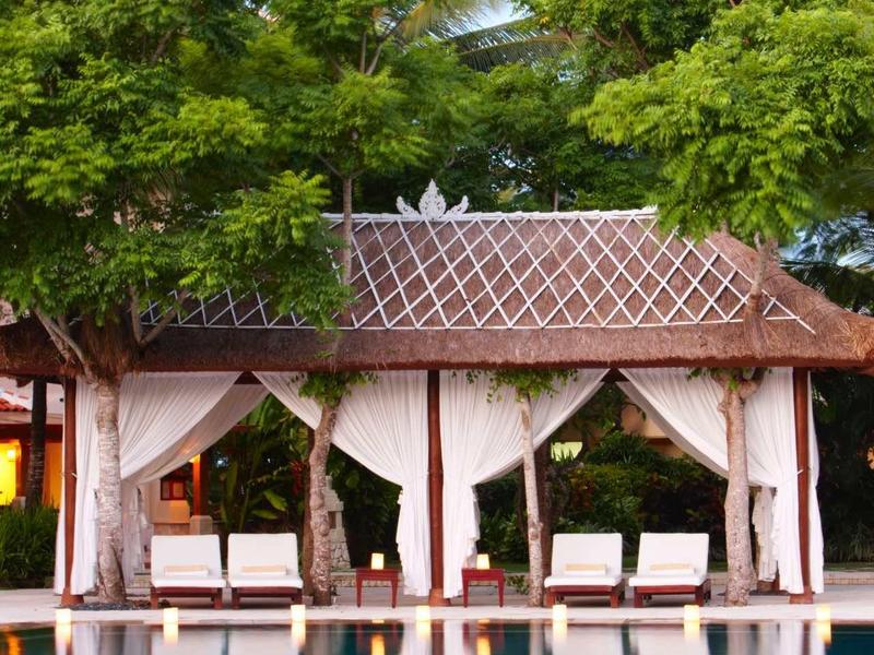 Piscina con pabellón y cortinas blancas, rodeada de árboles verdes y vegetación tropical.