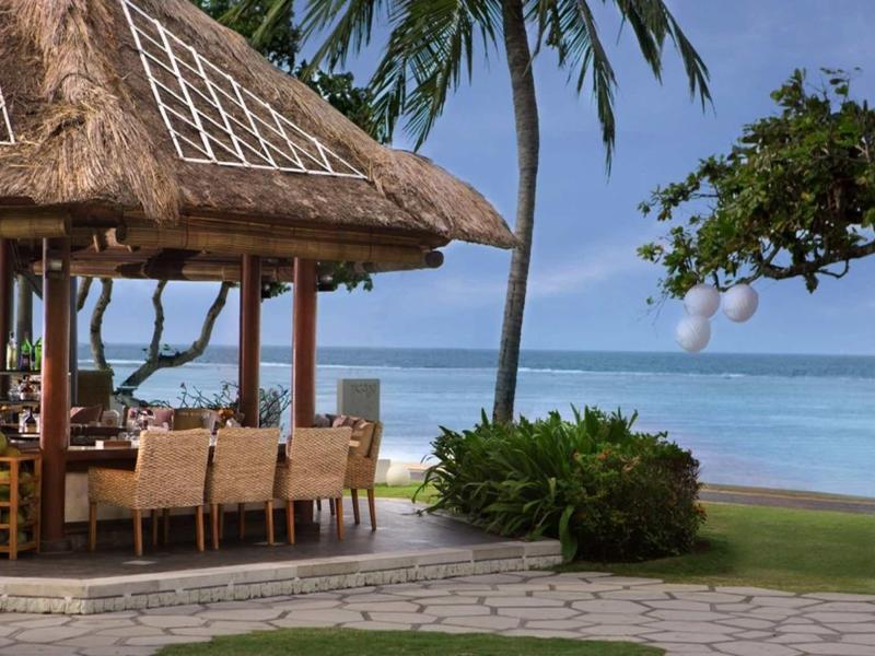 Restaurante abierto en la playa con mesas y sillas de madera, rodeado de palmas y vista al mar.