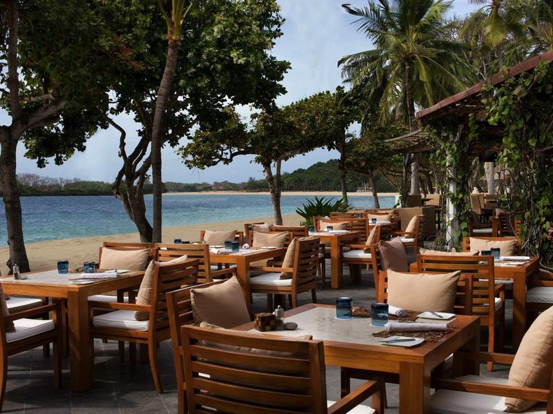 Área de comedor al aire libre con mesas y sillas de madera en la playa bajo los árboles.