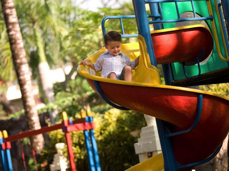 Un niño desliza por un tobogán en espiral colorido en un área exterior soleada con palmeras.