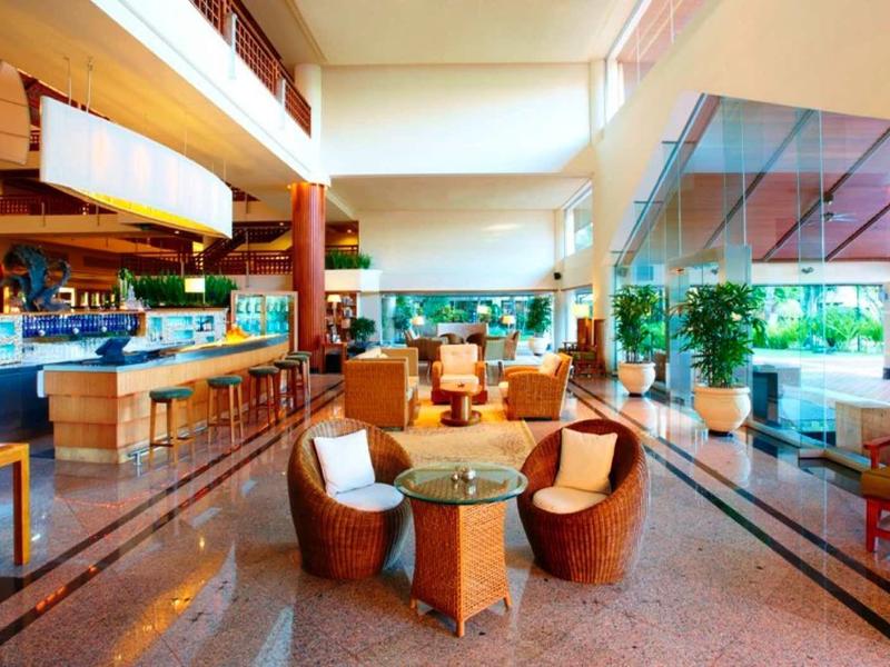 Área de salón de hotel luminosa con sillas modernas, mesas y grandes ventanales con mucha luz natural.