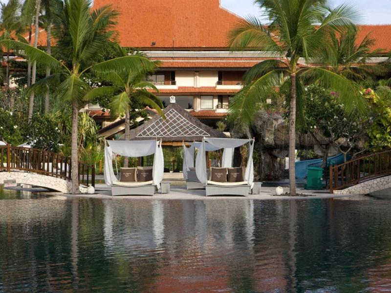 Gran piscina frente a hotel tropical con palmeras y sombrillas blancas.