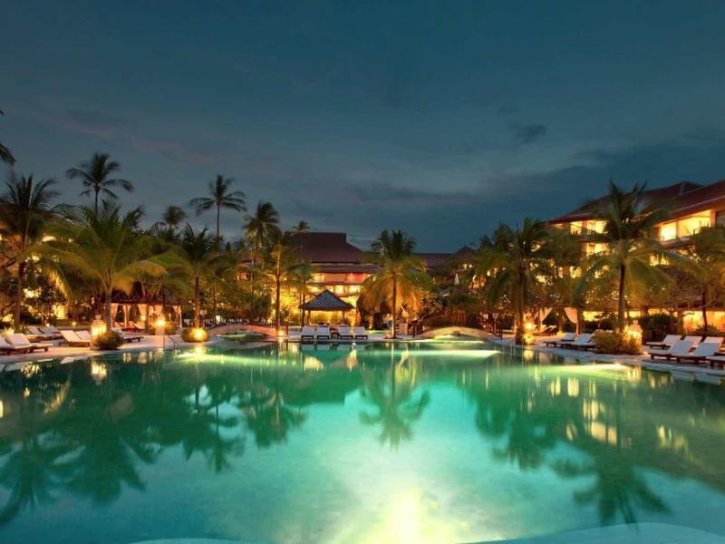 Gran piscina iluminada en un resort tropical al anochecer con palmeras y edificios
