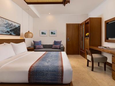 Helles Hotelzimmer mit großem Bett, Holzmöbeln und Sofa vor neutralen Wänden.