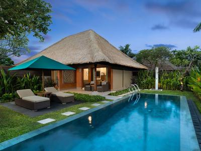 Ein Bungalow mit Reetdach neben einem rechteckigen Pool und Liegestühlen inmitten tropischer Pflanzen.