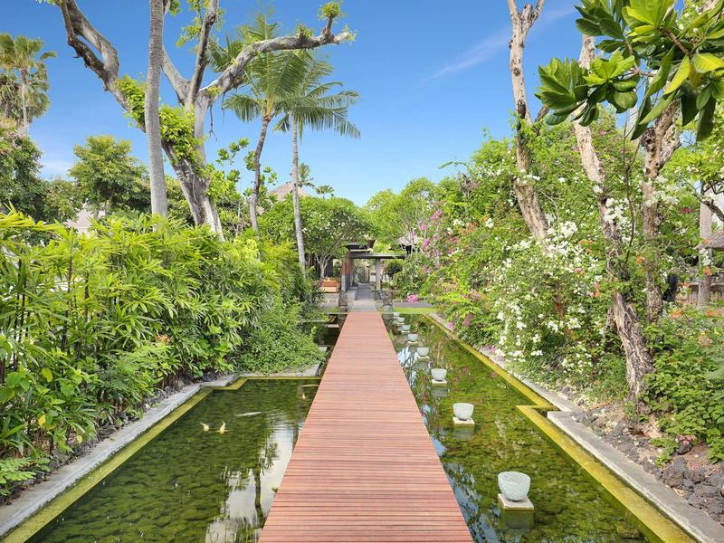 Een houten loopbrug loopt door een weelderige tropische tuin met waterbassins aan beide zijden.