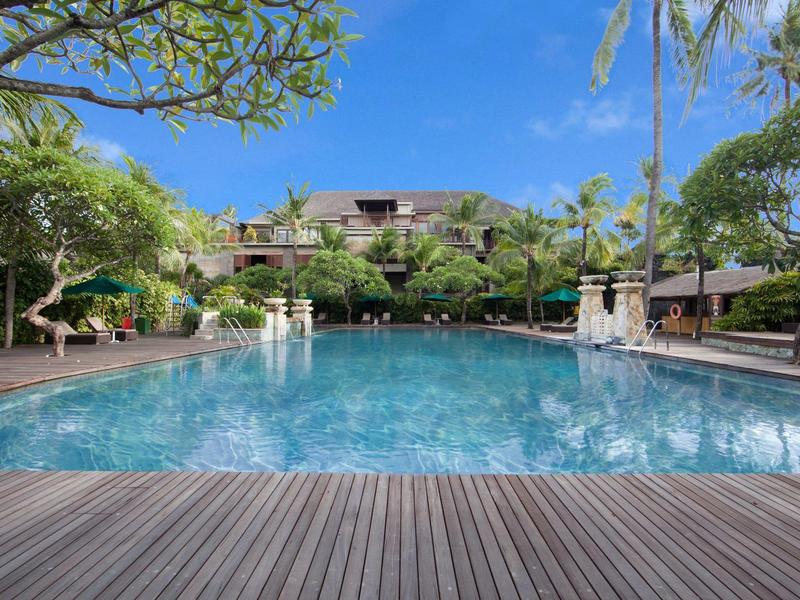 Groot zwembad met houten dek, omringd door palmbomen en ligstoelen onder blauwe lucht.