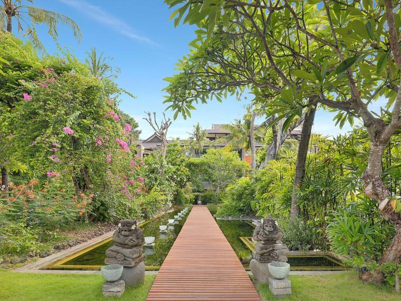 Houten pad door weelderige tuin naar luxe hotel onder blauwe lucht.