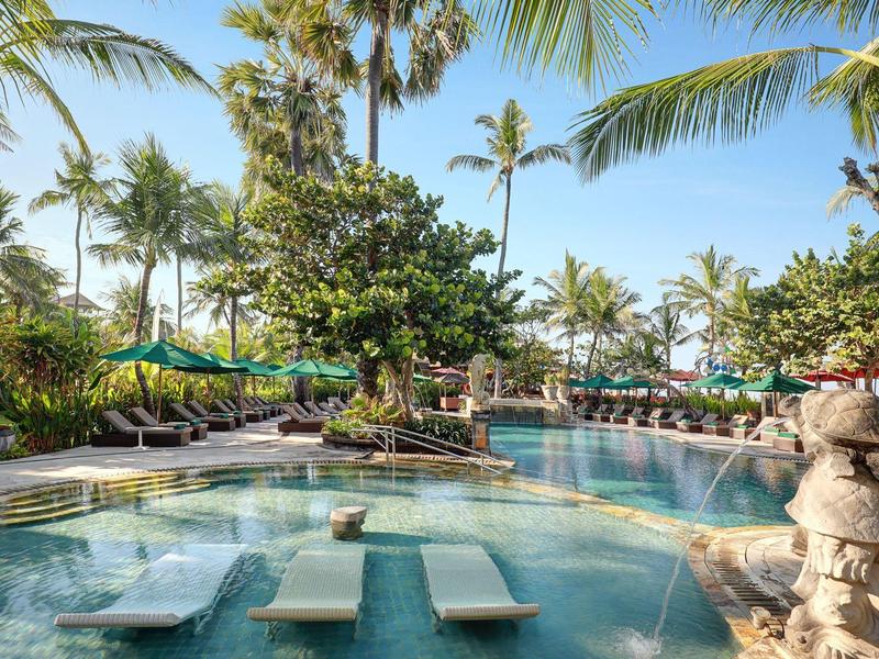 Een tropisch zwembadgebied met ligstoelen in het water en palmbomen op de achtergrond.