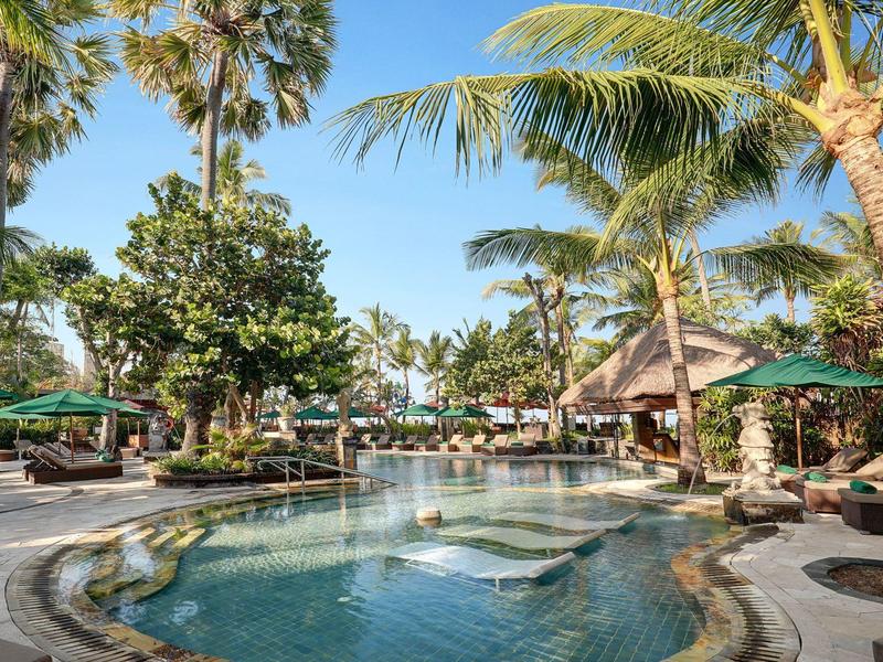 Tropisch hotelzwembad met palmbomen, ligstoelen en parasols onder een blauwe lucht.