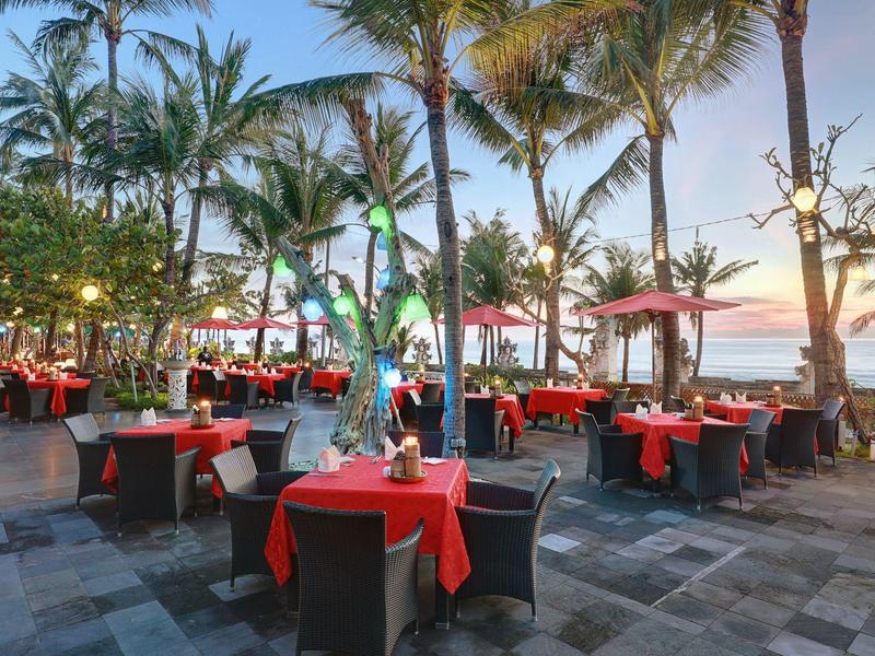 Buitenrestaurant met rode tafelkleden onder palmbomen op het strand bij zonsondergang.