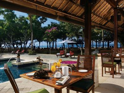 Área de restaurante al aire libre con sillas de madera y vista a la piscina y el mar bajo un techo de paja.