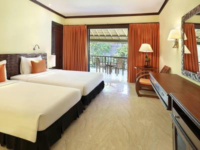 Habitación de hotel con dos camas individuales, balcón con vista a árboles, iluminación cálida y muebles de madera.