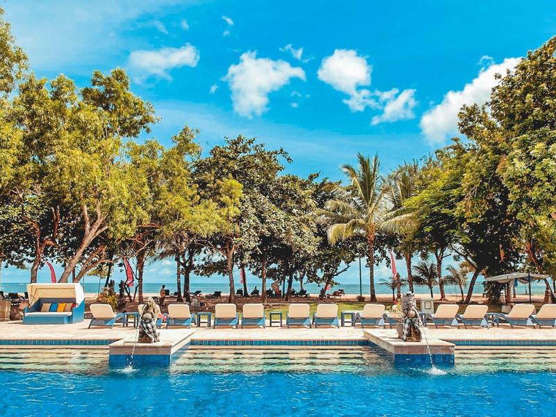 Piscina azul frente a una fila de tumbonas con árboles y mar al fondo en un día soleado.