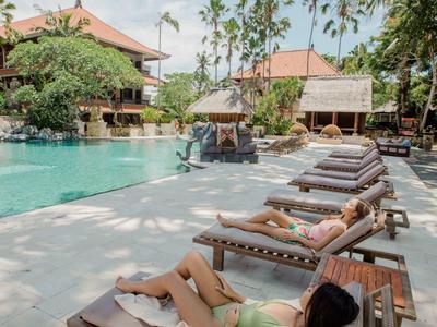 Frau entspannt auf Liegestuhl neben Pool, umgeben von Bäumen und Häusern bei sonnigem Wetter.