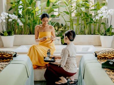 Une femme en robe jaune est assise sur un canapé tandis qu'une autre femme prépare un massage.