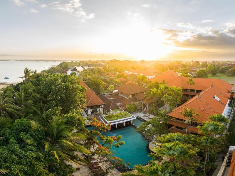 Luchtfoto van een resort met zwembad, omgeven door tropische planten en uitzicht op zee bij zonsondergang.