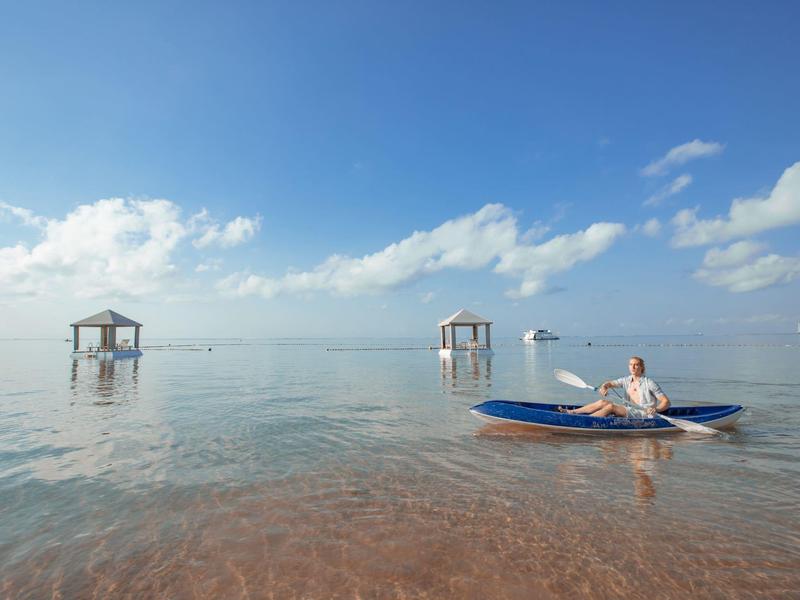 Vrouw zit in kajak op kalm water met paviljoens op de achtergrond onder een blauwe lucht.