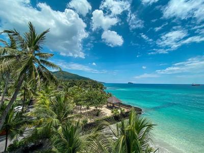 Klarer Himmel mit Wolken, türkisfarbenes Meer, Strände mit Palmen und grünen Hügeln im Hintergrund.