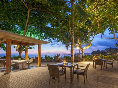 Terrasse mit Tischen und Stühlen, umgeben von Bäumen, Blick auf Wasser beim Sonnenuntergang.