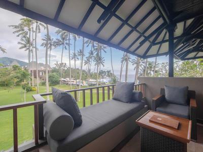 Überdachte Terrasse mit grauer Sitzbank, Kissen, Holztisch und Blick auf Palmen und Grünfläche.