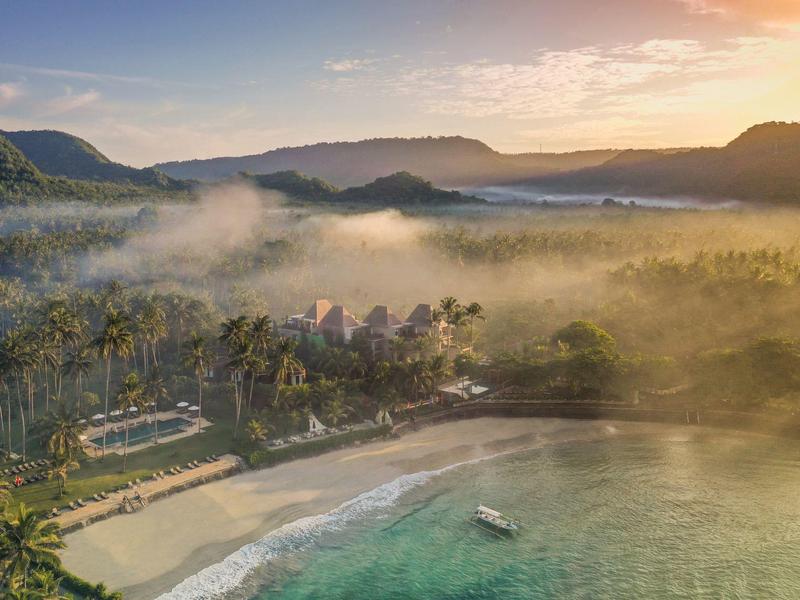 Küstenlandschaft mit Strand, grünem Wald, Boot auf türkisblauem Wasser und Bergen im Hintergrund bei Sonnenaufgang.