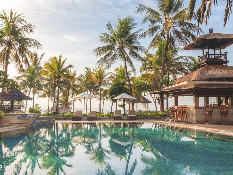 Ein ruhiger Pool mit klar reflektierendem Wasser, umgeben von Palmen und Sonnenschirmen bei Sonnenschein.