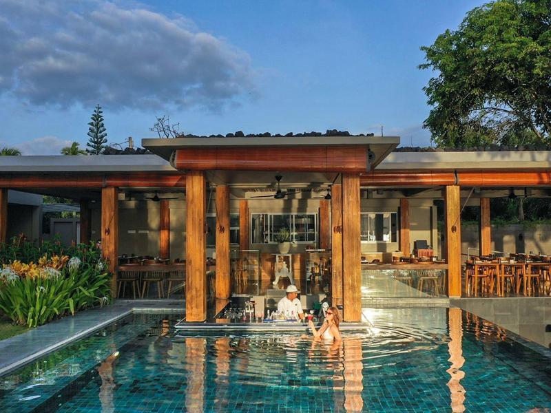 Moderner Holzbau mit großem Pool und Sitzbereich, umgeben von grünen Bäumen und blauem Himmel.