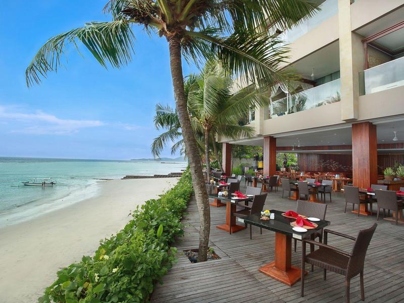 Strandrestaurant mit Holzboden, Tischsets und Stühlen neben Palmen und weißem Sandstrand.