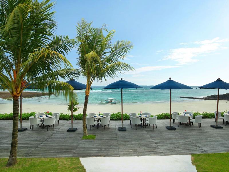 Strandpromenade mit Sonnenschirmen, Sitzgruppen und Palmen vor ruhigem Meer und blauem Himmel.