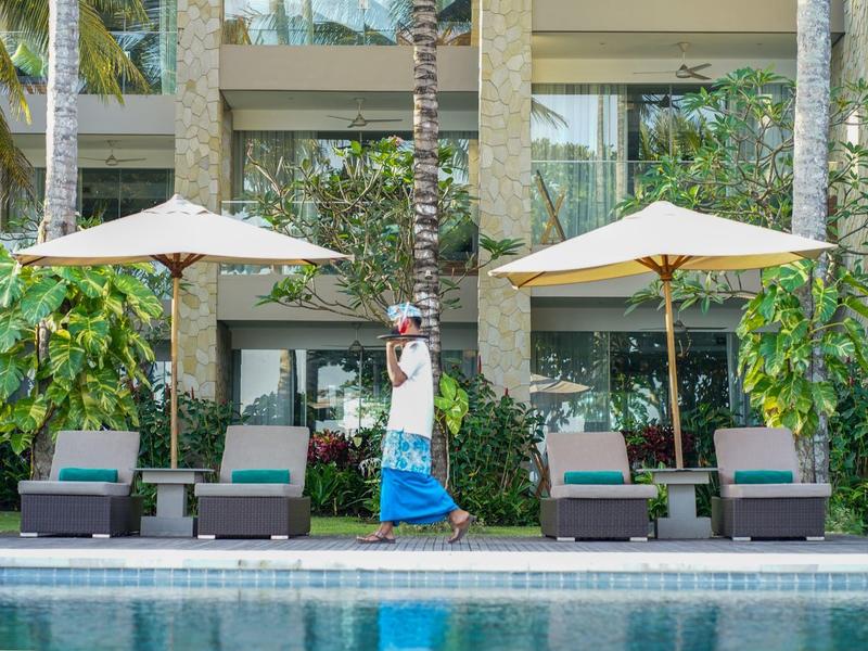 Außenpool mit Liegen, Sonnenschirmen, Palmen und Person im blauen Rock vor Glasgebäude.