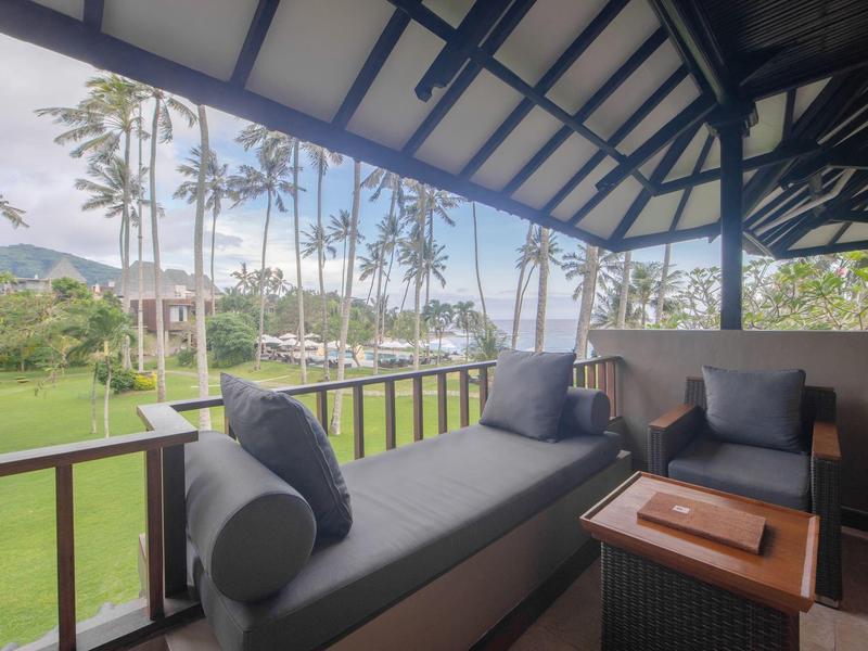 Überdachte Terrasse mit grauer Sitzbank, Kissen, Holztisch und Blick auf Palmen und Grünfläche.
