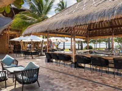 Covered beach bar with wooden chairs and tables, palm trees, and sea view in the background.