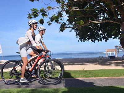 Two cyclists wearing helmets ride along the beach under a tree on a clear day.