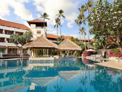 Hotel with large pool, palm trees, and sun umbrellas in a tropical garden under blue sky.