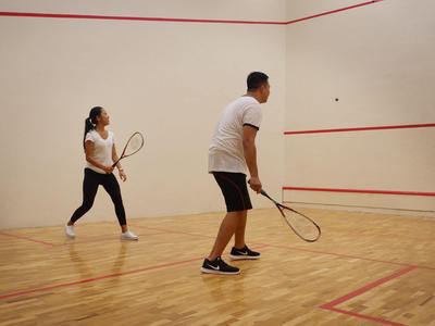Two people playing squash in a bright, empty squash court with a wooden floor.