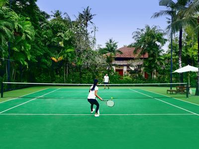 Two people playing tennis on a green court surrounded by palm trees and a building in the background.