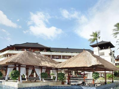 Zwembadgebied met rieten paviljoenen en ligstoelen voor een hotel onder een blauwe lucht.