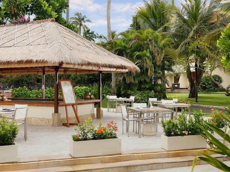Open zitgedeelte met tafels en stoelen naast een paviljoen met rieten dak in een tropische tuin.