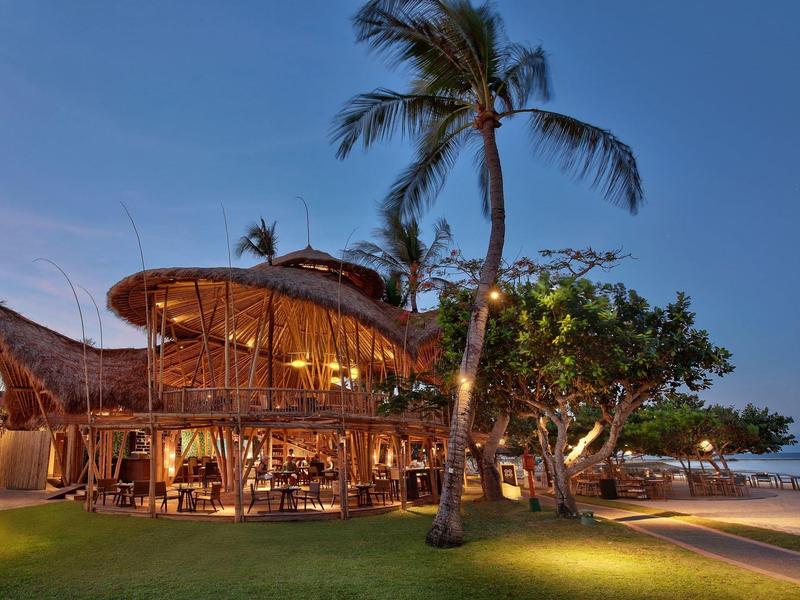 Modern restaurant met houten structuur en palmbomen aan het strand bij schemering.