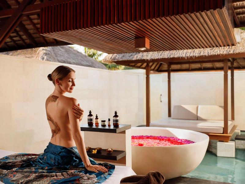 Vrouw zit naast badkuip vol rozenblaadjes onder open dak in ontspannende spa omgeving.