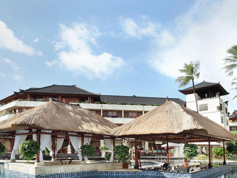 Zwembadgebied met rieten paviljoenen en ligstoelen voor een hotel onder een blauwe lucht.