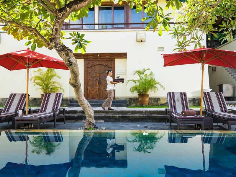 Zwembad met ligstoelen en rode parasols voor een gebouw met een sierlijke houten deur.