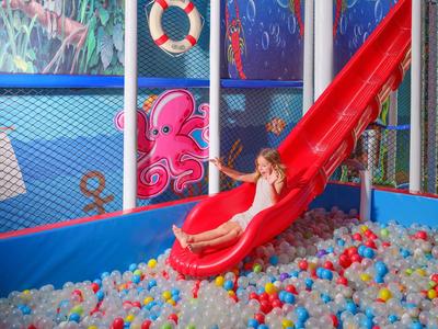 Kind rutscht eine rote Rutsche hinunter in ein Bällebad im Indoor-Spielbereich.