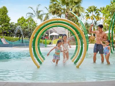 Familie spielt im flachen Pool mit Wasserspielen und grünem Hintergrund.
