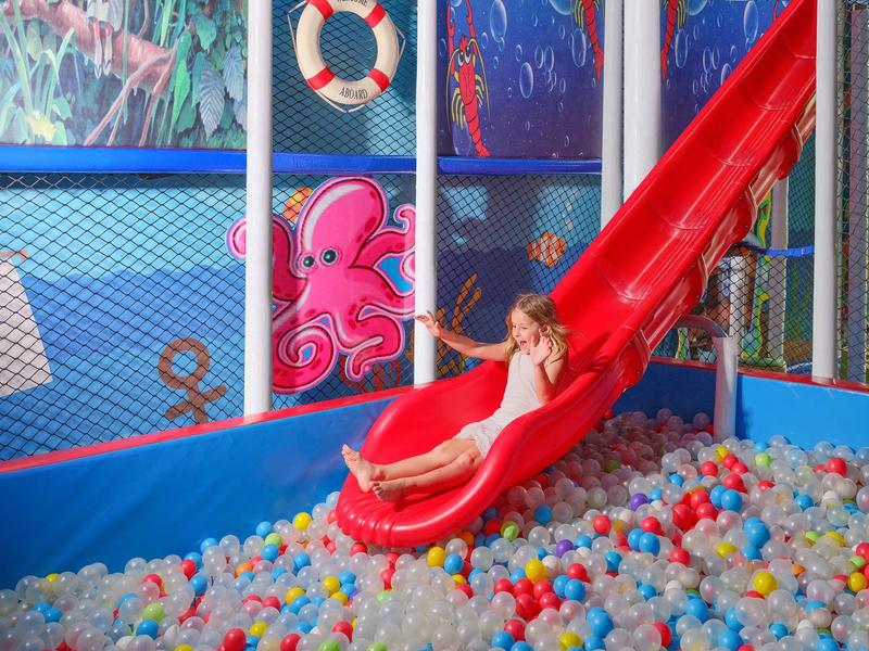 Kind rutscht eine rote Rutsche hinunter in ein Bällebad im Indoor-Spielbereich.
