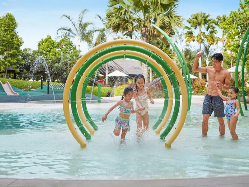 Familie spielt im flachen Pool mit Wasserspielen und grünem Hintergrund.