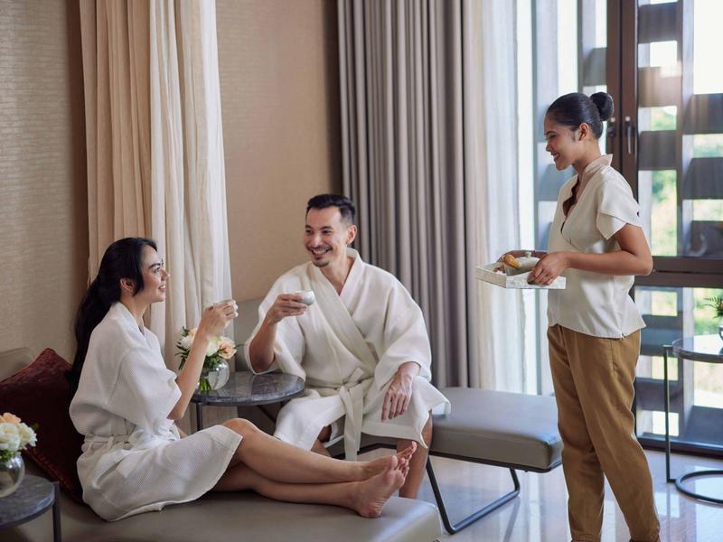 Paar in Bademänteln entspannt sich im Hotelzimmer, Kellner serviert Getränke.