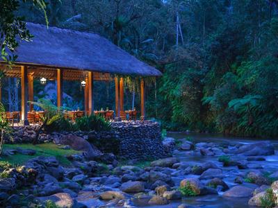 Cozy riverside thatched pavilion lit warmly at dusk surrounded by lush forest and rocks.